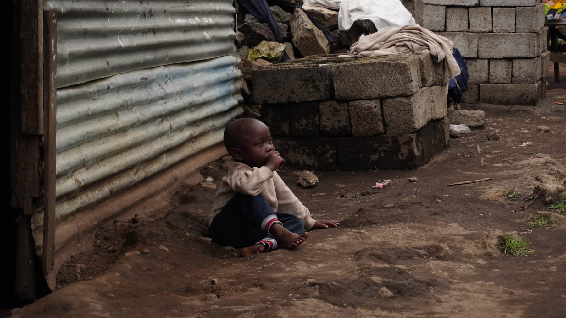 A young boy sits alone on the ground beside a corrugated metal shack in eastern DRC