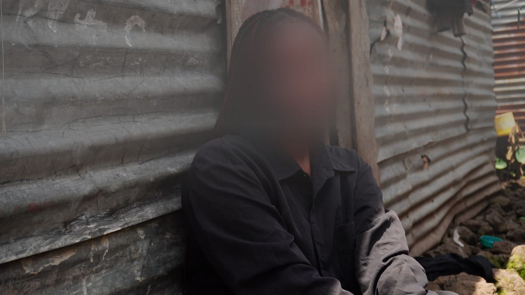 En kvinne i Øst-Kongo sitter mot en bølgeblikkvegg, med ansiktet i dyp skygge for å beskytte identiteten hennes